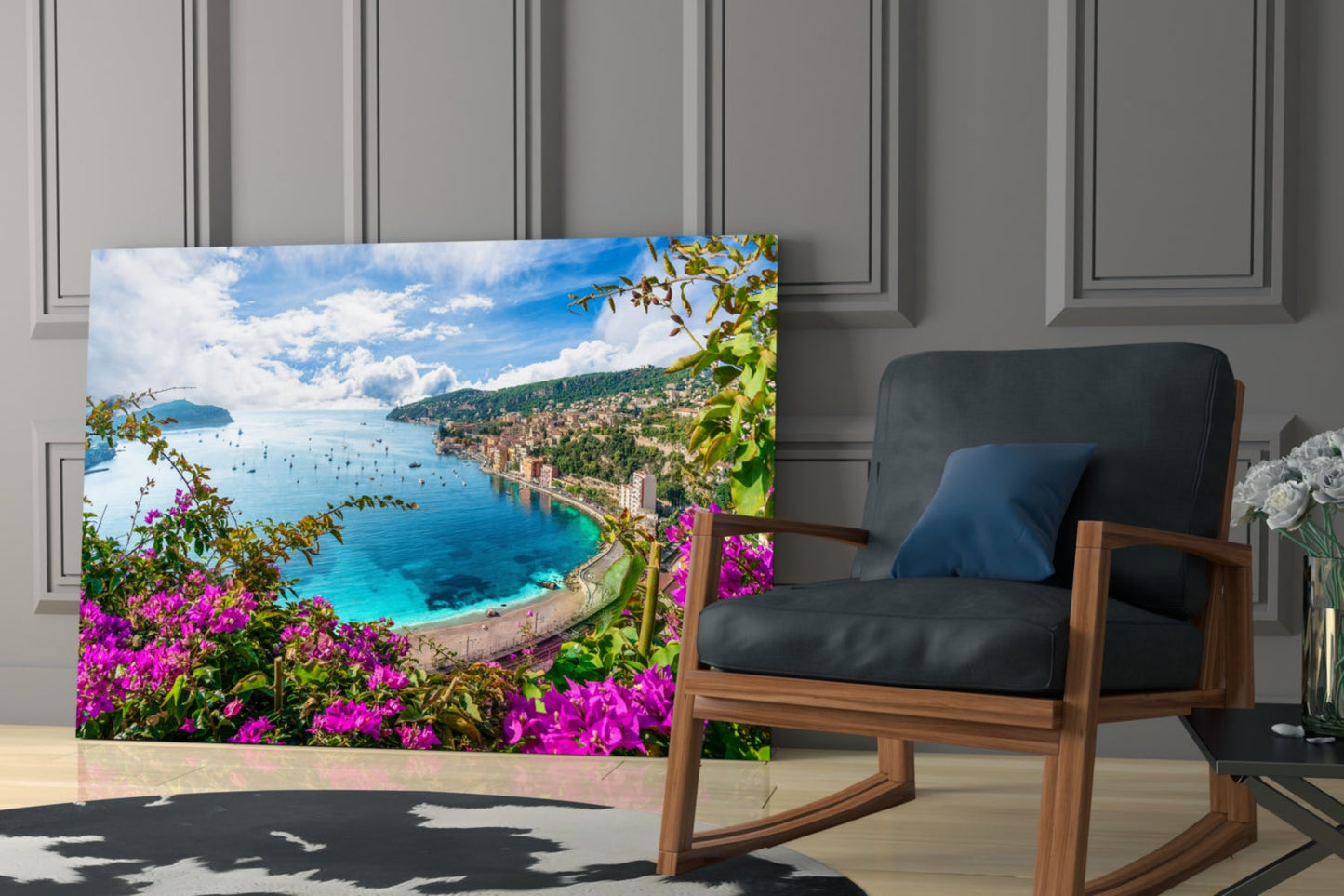 Glass wall art displaying a coastal Mediterranean landscape with magenta flowers, turquoise sea, and hillside buildings, positioned beside a wooden chair in a modern interior.