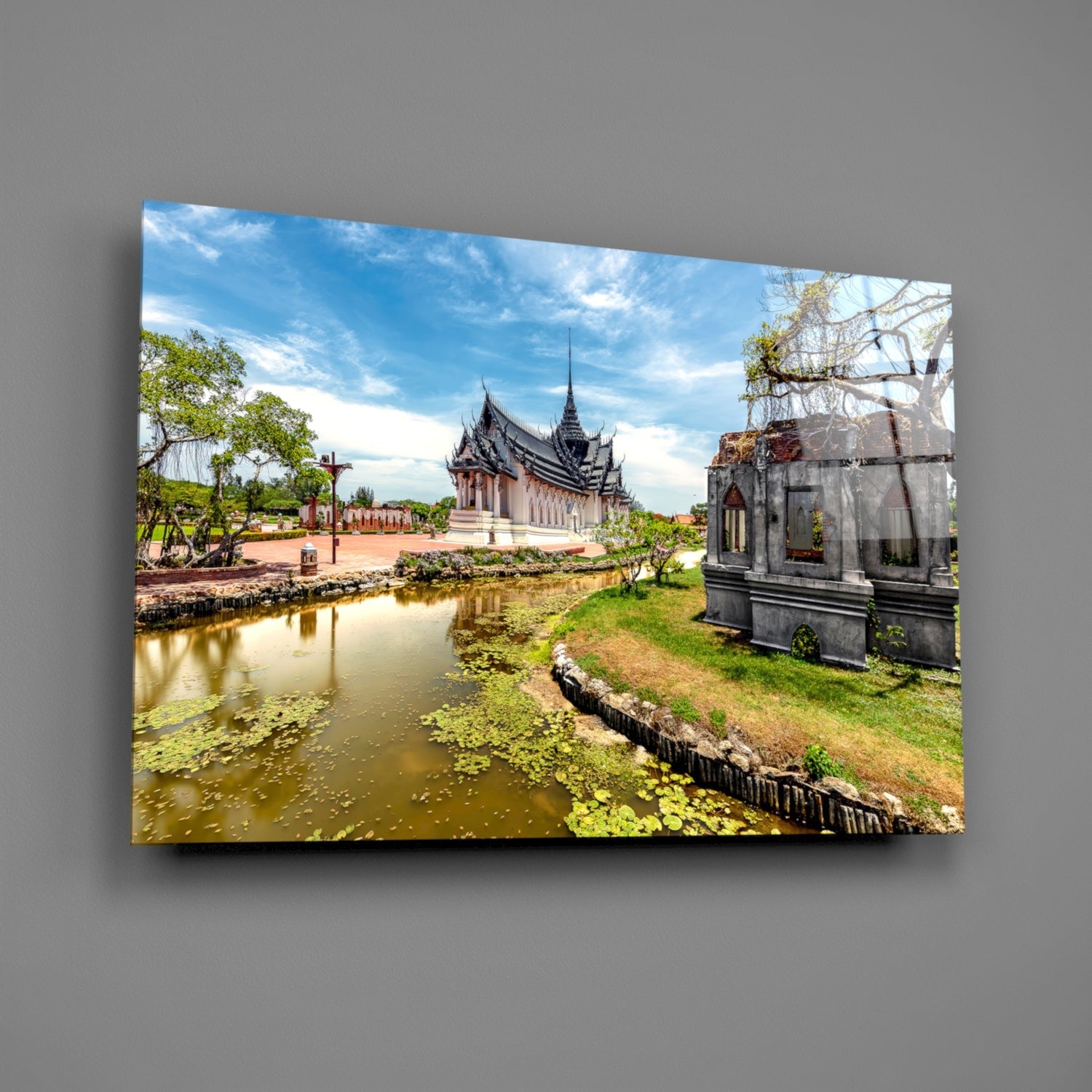 Glass wall art displaying a Thai temple with distinctive black spires beside a calm pond with lily pads and green landscaping under blue sky.
