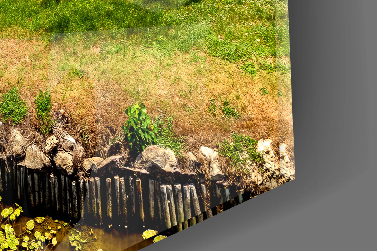 Aerial view of a hillside landscape with green grass, brown earth, wooden fence, rocks, and a small plant, printed on glass wall art.