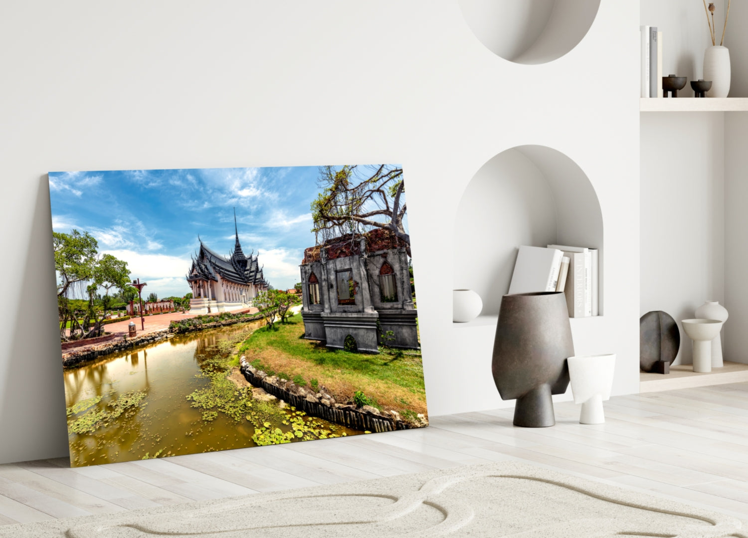 Glass wall art leaning against white wall displaying Thai temple by water with blue sky and green landscape.