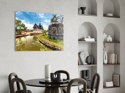 Glass wall art displaying a Thai temple beside a waterway with blue sky, hung above a dining table in a modern interior.
