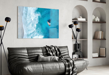 Oceanside Glass Wall Art displaying aerial turquoise ocean waves and a small boat, mounted above a black leather sofa in a modern living room.