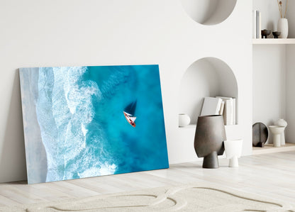 Aerial view glass art leaning against white wall showing turquoise ocean waves and red sailboat.