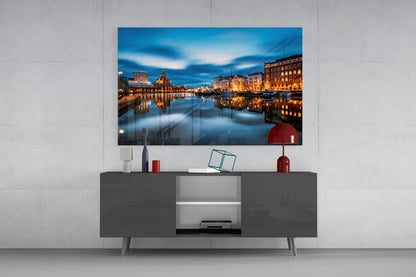 Glass wall art displaying a twilight cityscape with illuminated buildings reflected in calm water, mounted above a dark TV console in a modern living room setting.