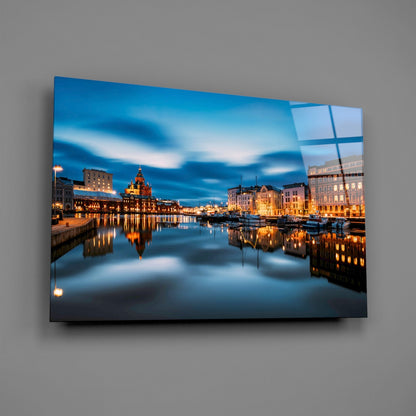 Glass wall art mounted on gray wall showing illuminated Helsinki waterfront at twilight with reflecting buildings and boats.