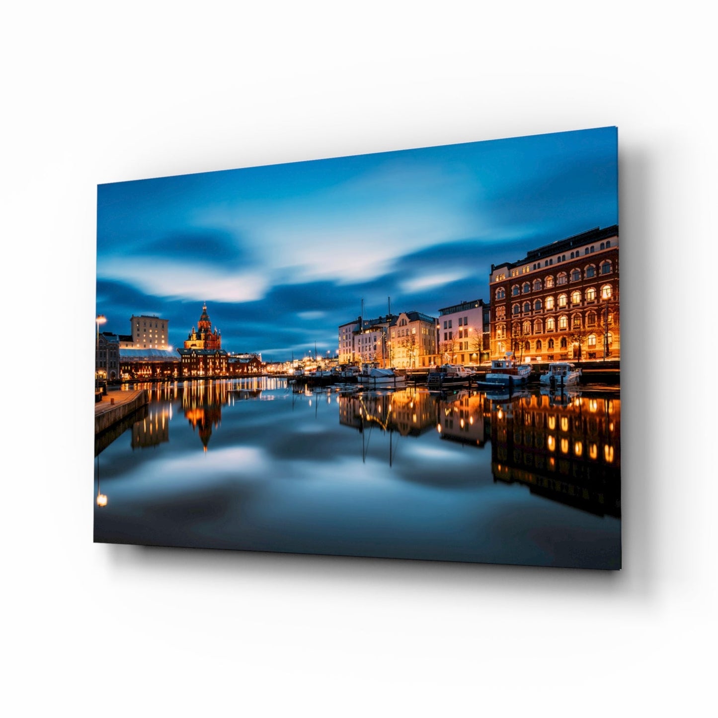 Glass wall art print displayed at an angle showing a twilight cityscape with illuminated waterfront buildings reflected in still water.