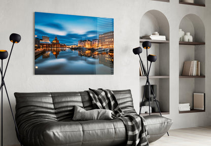 Glass wall art displaying illuminated Helsinki waterfront buildings reflected in calm blue water, hung above gray leather sofa.