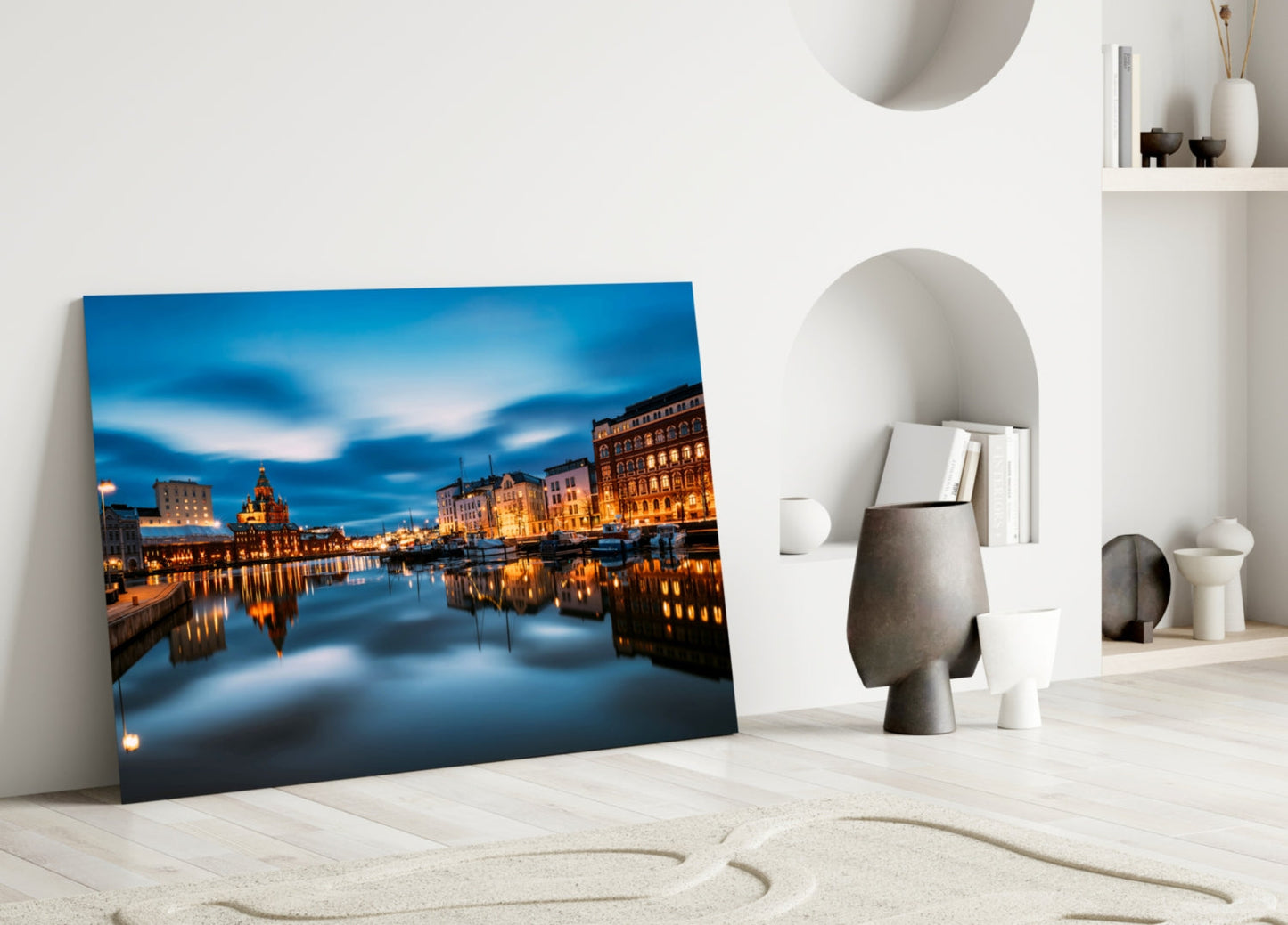 Glass wall art leaning against white wall displaying illuminated Helsinki cityscape with blue evening sky and golden building reflections.
