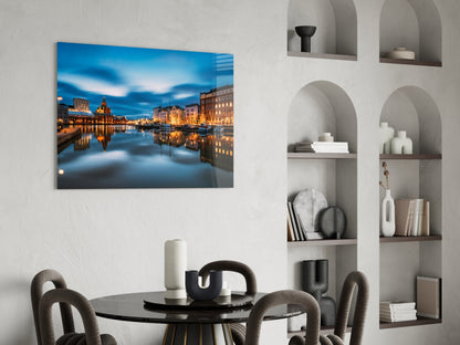 Glass wall art depicting an illuminated European waterfront cityscape at twilight, displayed above a dining table in a modern interior.