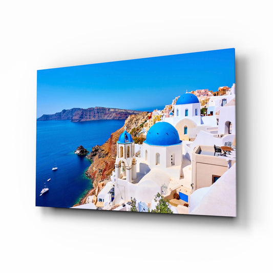 Glass wall art print mounted at an angle displaying a white-walled church with blue dome overlooking a blue Aegean Sea and distant cliffs.