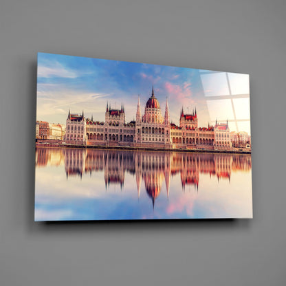 Glass wall art displaying the Hungarian Parliament Building reflected in still water at sunset, mounted on gray wall.