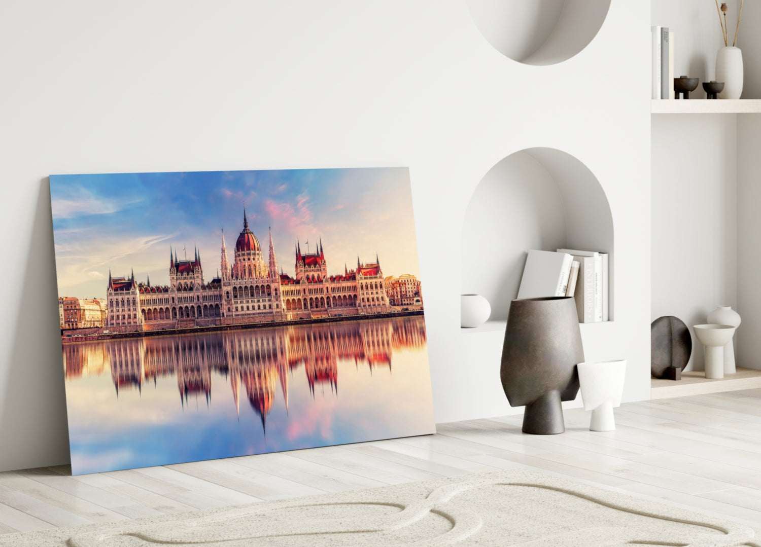 Glass wall art leaning against white wall displays Budapest Parliament Building at sunset reflected in water, with warm pink and blue tones.