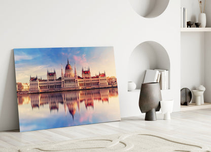 Glass wall art leaning against white wall displays Budapest Parliament Building at sunset reflected in water, with warm pink and blue tones.