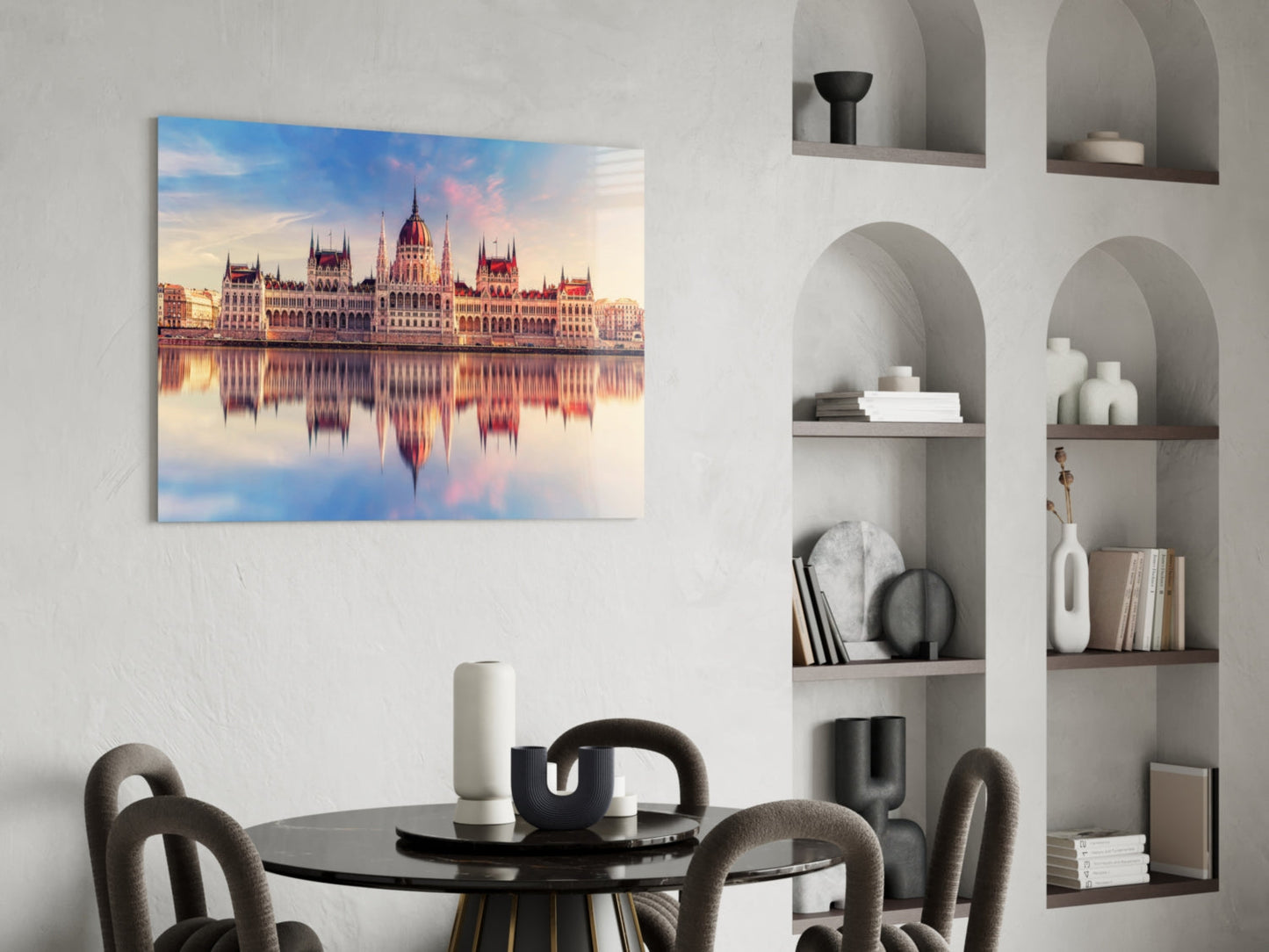 Glass wall art print of Hungarian Parliament Building reflected in water, displayed above round black dining table in modern living room with built-in shelving.