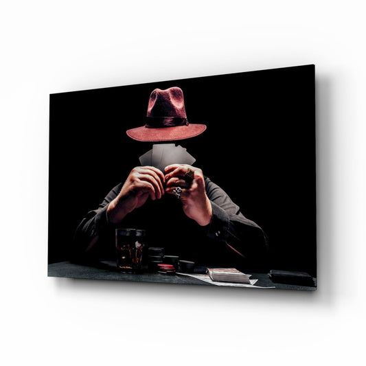 Glass wall art showing a headless figure in dark jacket wearing a pink fedora hat, hands positioned over playing cards and poker chips on a black surface against black background.