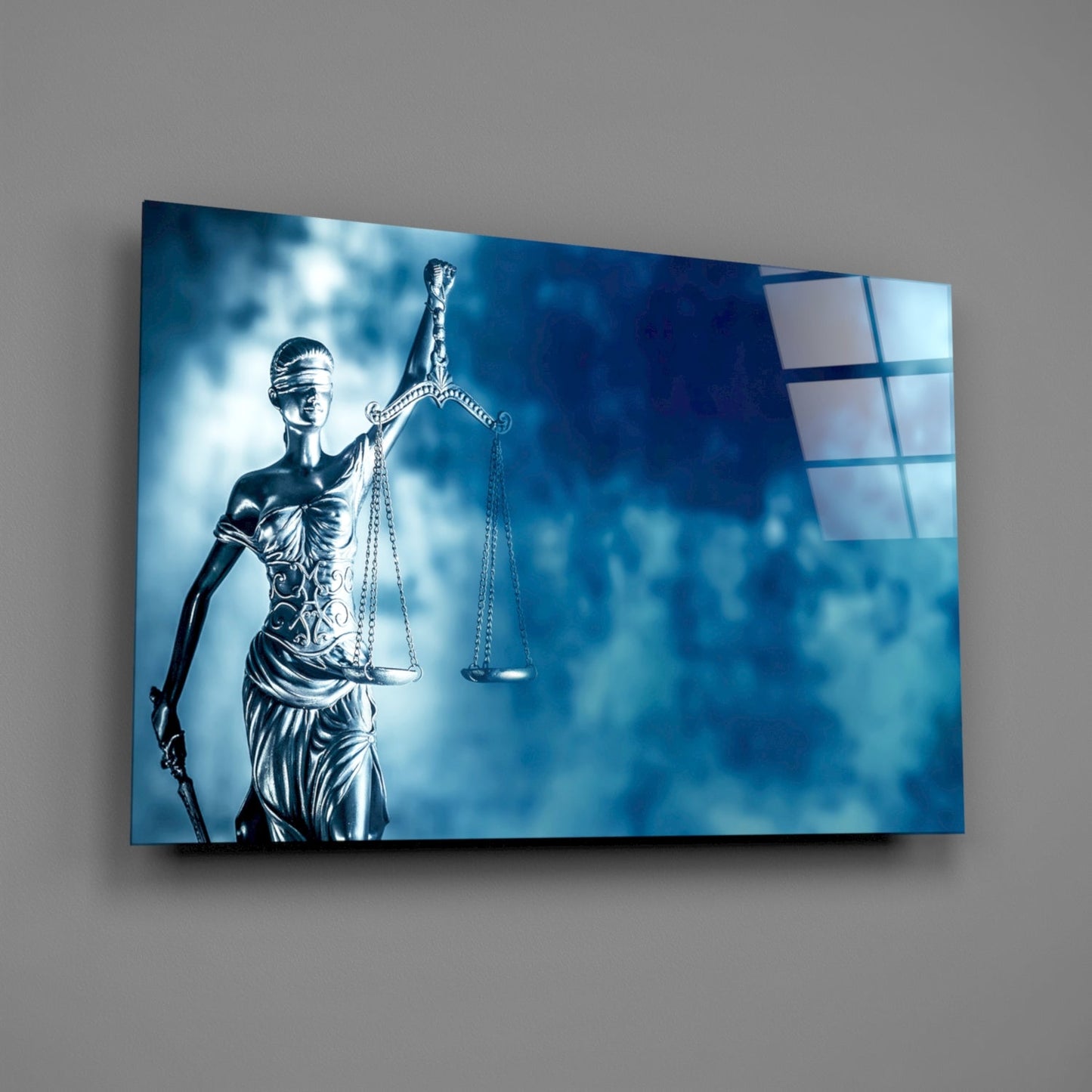 Angled view of a glass wall art panel displaying Lady Justice statue in silver against a blue cloudy background, mounted on gray wall.