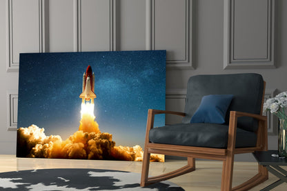 Glass wall art displaying a space shuttle launching through orange clouds against a starry blue sky, installed in a modern living room beside a wooden chair.