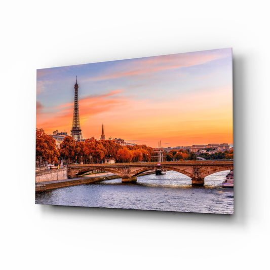 Glass wall art panel displaying Paris Seine river with stone bridge, Eiffel Tower, autumn trees, and vibrant orange sunset sky.