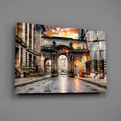 George Square Glass Wall Art mounted on gray wall, showing historic stone archway at sunset with wet pavement and buildings.