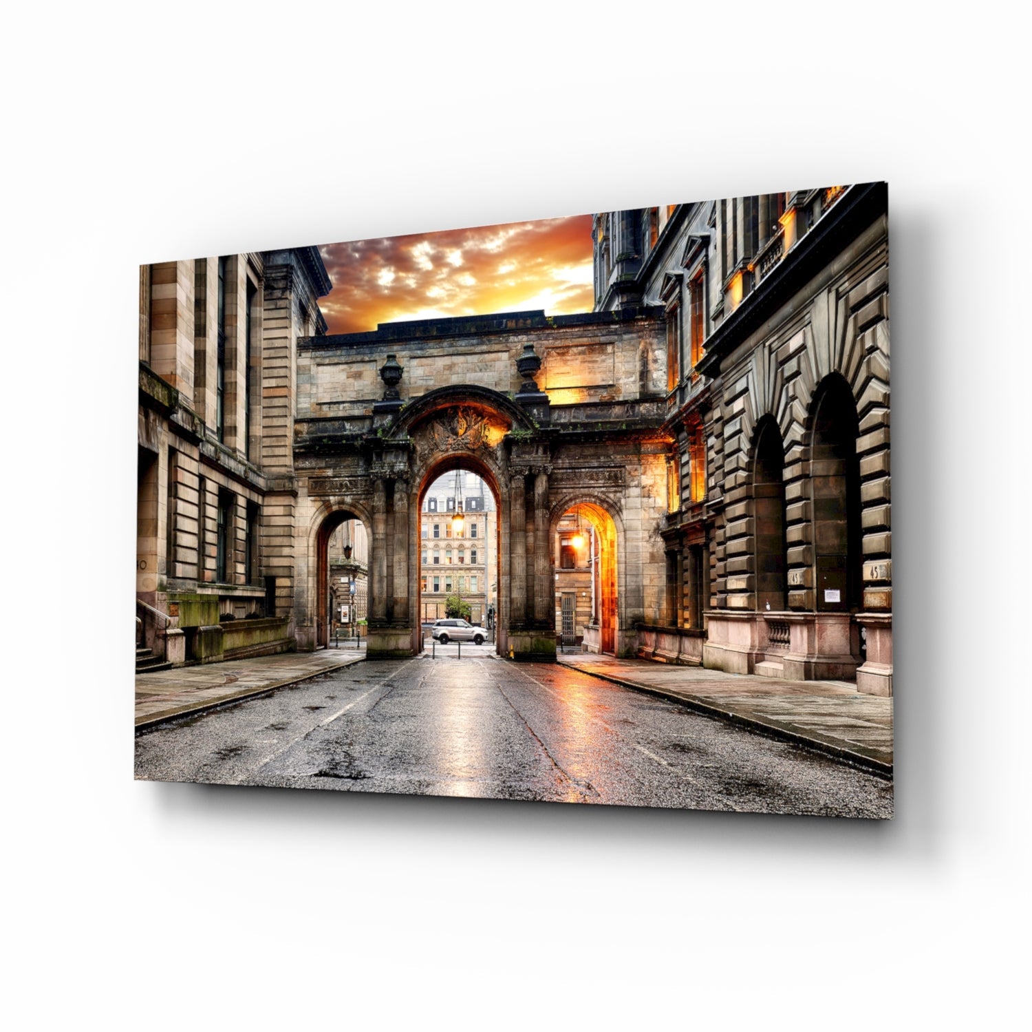 Glass wall art print displayed at an angle showing a historic stone archway at sunset with warm golden light reflecting off wet pavement.