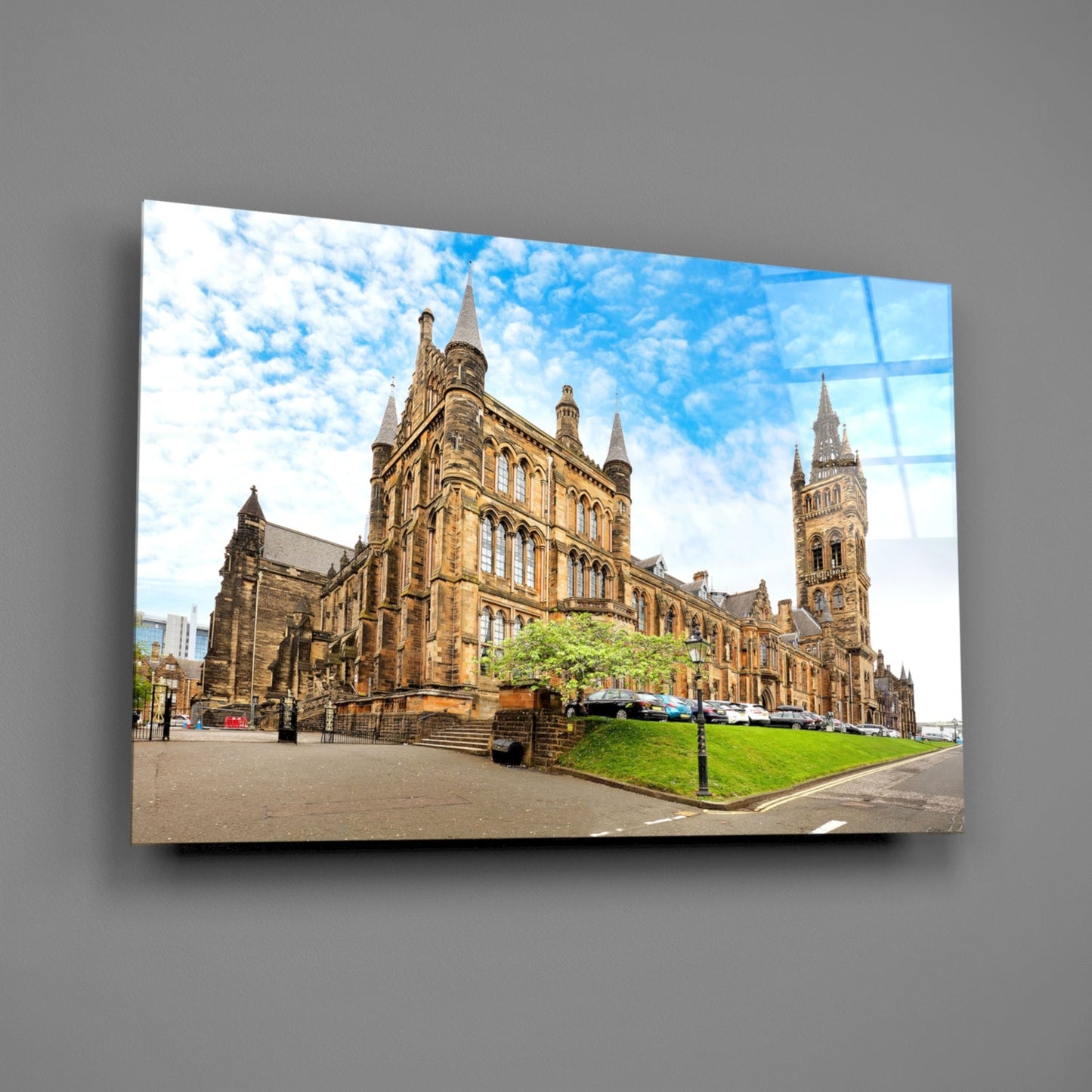 Glass wall art panel displaying University of Glasgow's Gothic stone buildings with green lawn and blue sky, mounted on gray wall.