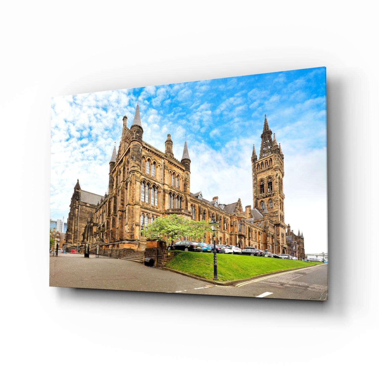 Glass wall art print of University of Glasgow's Gothic sandstone buildings with green lawn and blue sky, displayed at an angle showing depth.
