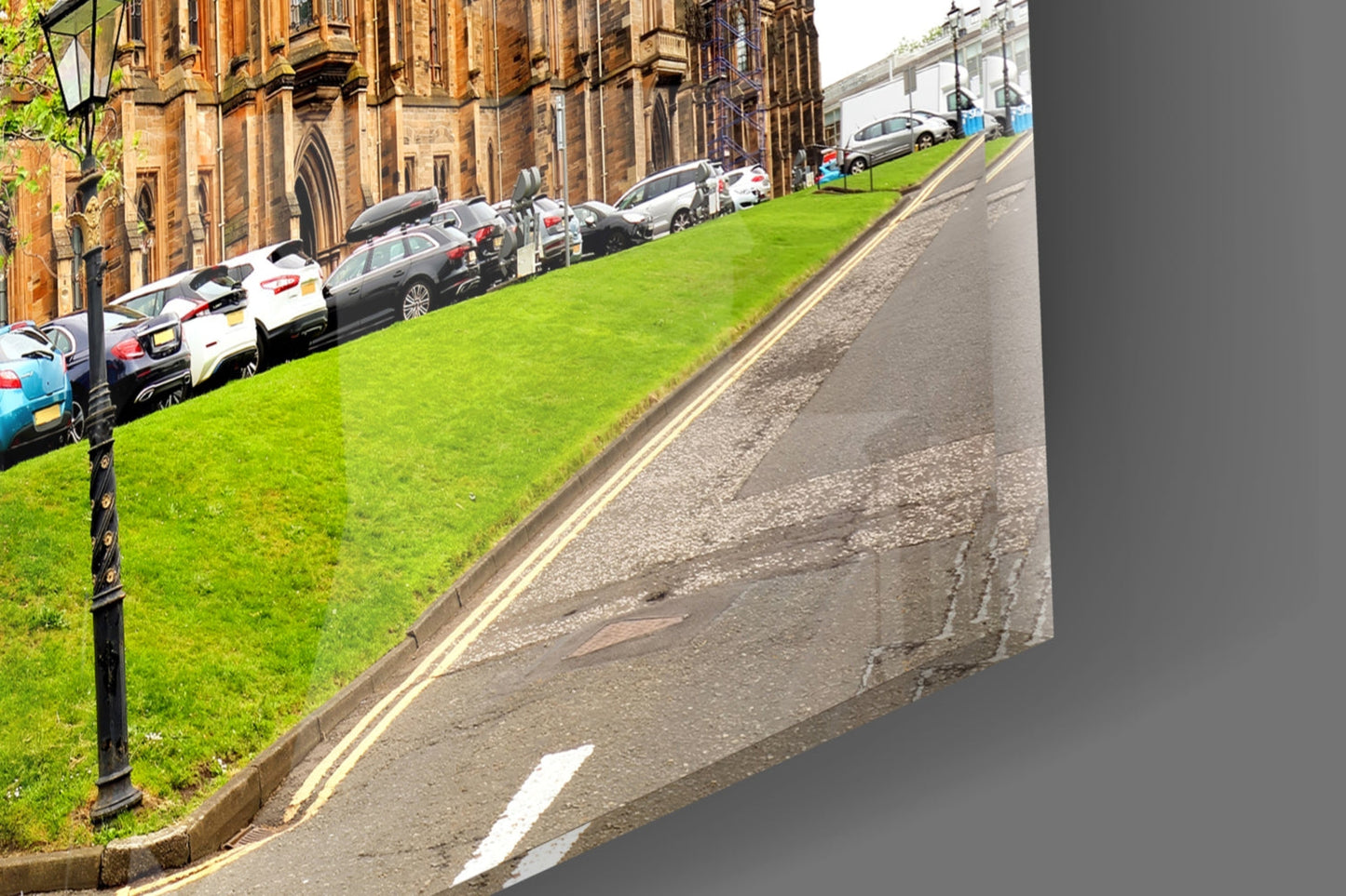 Sloped grassy hillside with parked cars beside historic stone building and street, taken from elevated angle.
