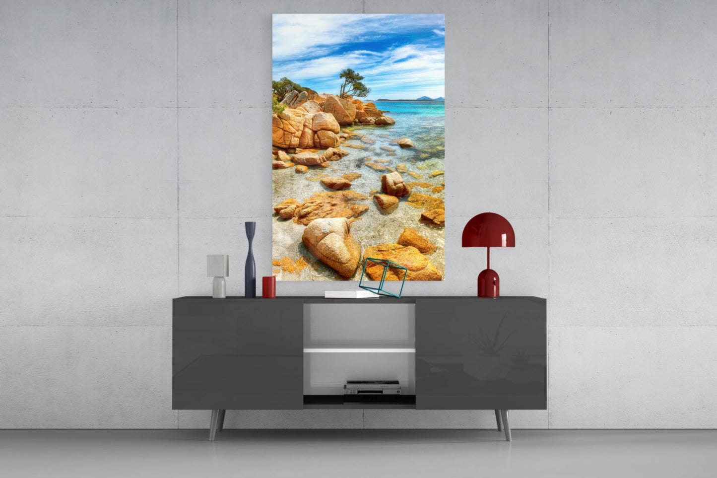 Beach wall art glass panel mounted above dark gray media console, displaying rocky coastline with turquoise water and blue sky.