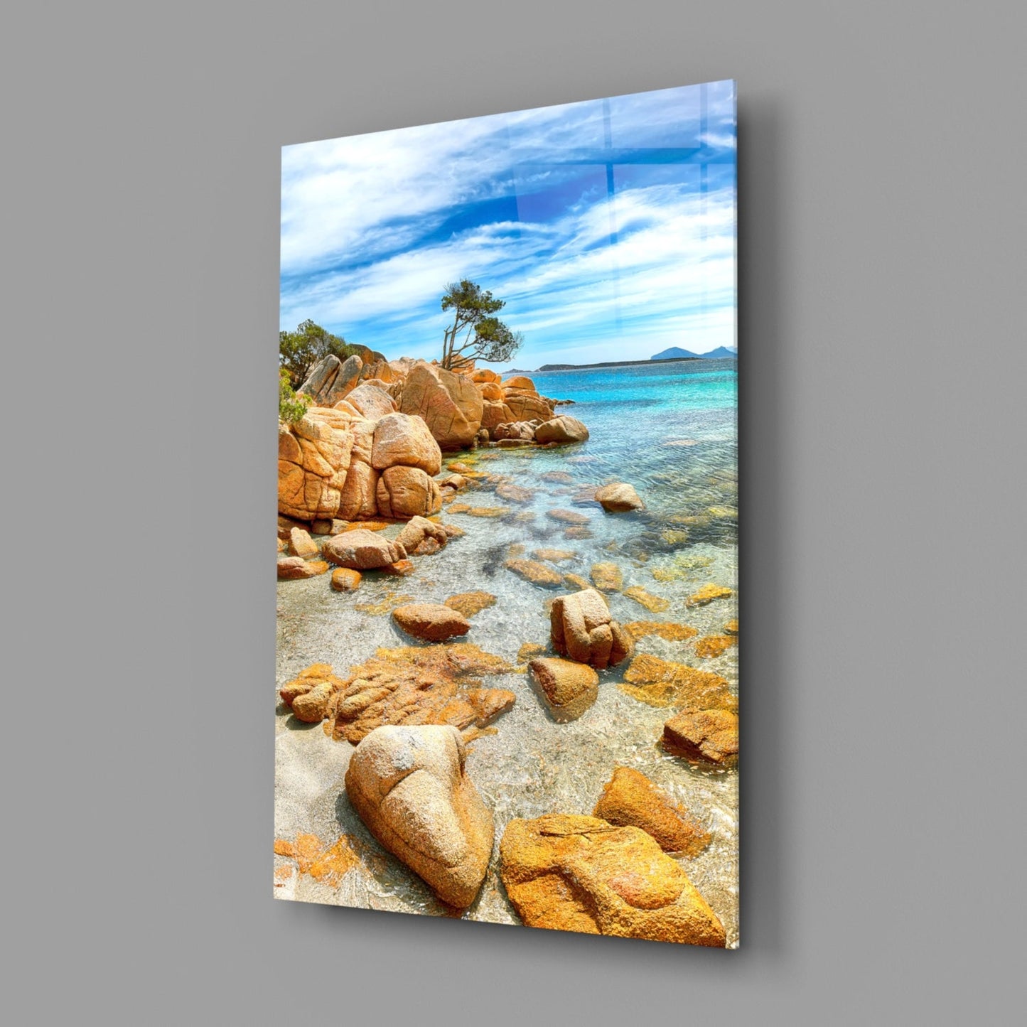 Glass wall art displaying a rocky Mediterranean beach with turquoise water and distant mountains under blue sky.