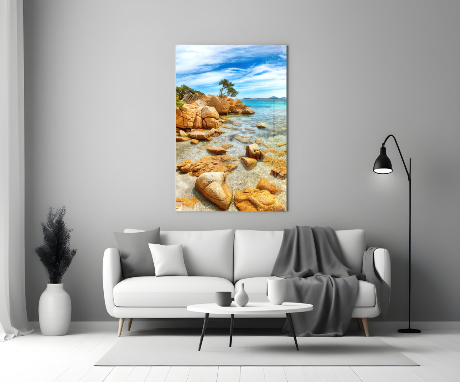 Beach View Glass Wall Art displayed above white sofa showing rocky coastline with turquoise water and blue sky.