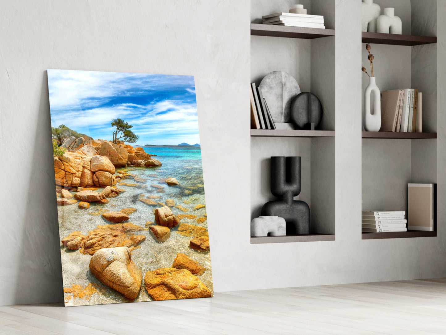 Glass wall art leaning against white wall displays rocky beach with golden boulders, turquoise water, and blue sky.