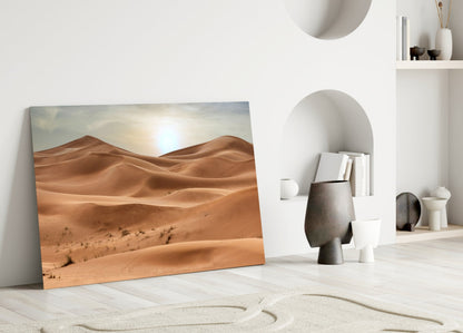 Desert landscape glass wall art leaning against white wall in minimalist room with decorative objects.