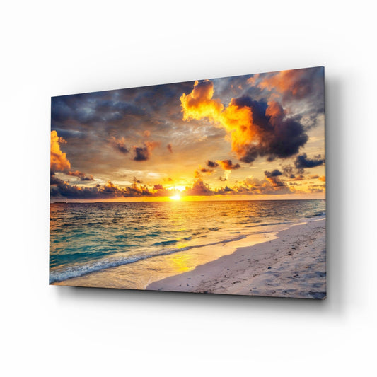 Angled view of glass wall art depicting a golden sunset over a beach with dramatic orange and dark clouds reflected on calm turquoise water.