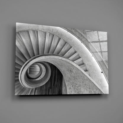 Monochrome photograph of a spiral staircase viewed from above, showing concentric circular steps and concrete walls descending into darkness.