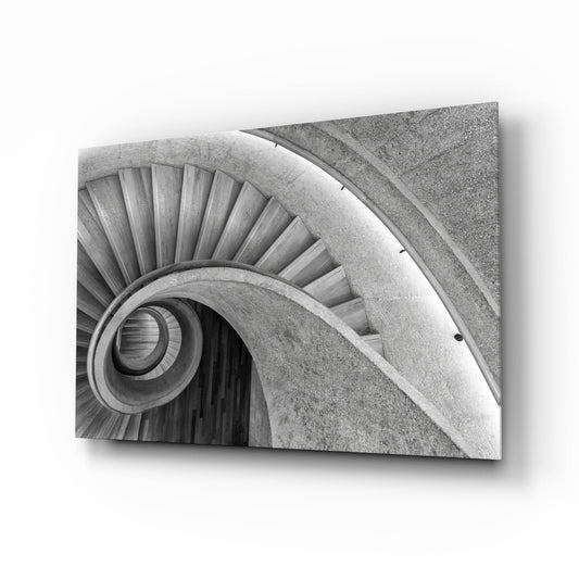 Black and white photograph of a spiral staircase viewed from above, showing concentric circular steps and handrails in concrete.