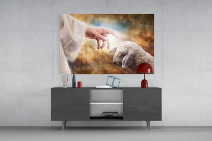 Glass wall art depicting a divine hand reaching toward a white lamb against a golden sky, mounted above a dark media console.