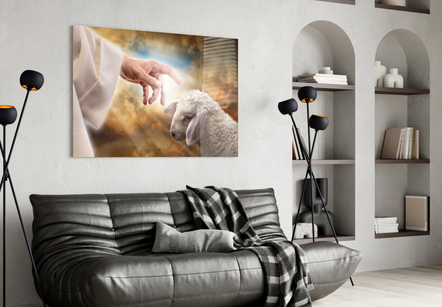Glass wall art depicting a hand reaching toward a white lamb against a golden, cloudy background, displayed above a black leather sofa in a modern living room.