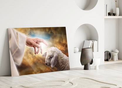 Glass wall art leaning against white wall showing a hand reaching toward a white sheep against golden cloudy sky background, displayed in minimalist interior setting.