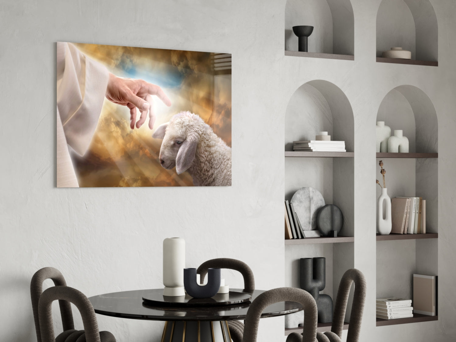 Glass wall art depicting a hand reaching toward a white lamb against golden clouds, displayed above a black dining table in a modern interior.