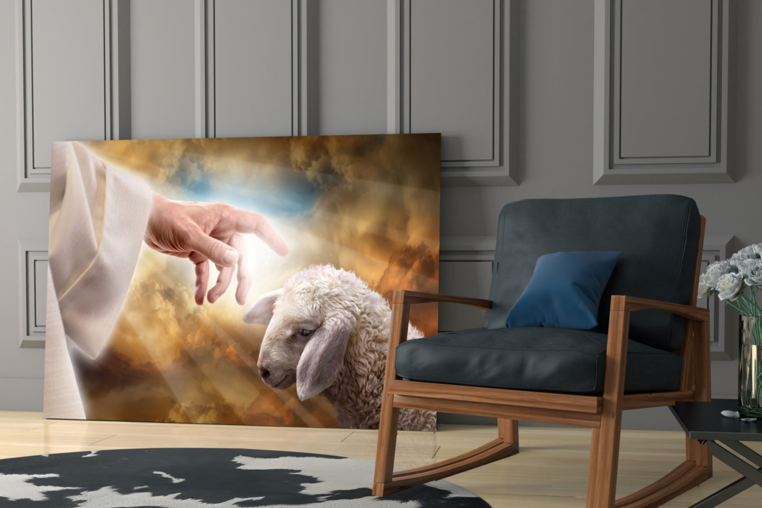 Glass wall art depicting a divine hand reaching toward a white lamb against golden clouds, displayed in a modern interior with wooden chair.