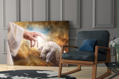 Glass wall art depicting a divine hand reaching toward a white lamb against golden clouds, displayed in a modern interior with wooden chair.