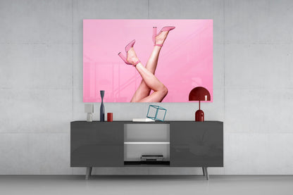 Pink glass wall art depicting legs in heels displayed above dark media console in modern interior setting.