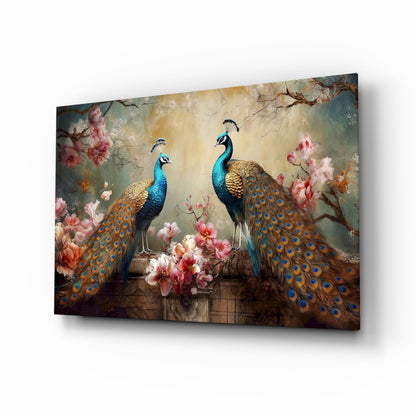 Angled three-quarter view of glass wall art featuring three blue-headed peacocks with golden tail feathers surrounded by pink flowers against a muted background.