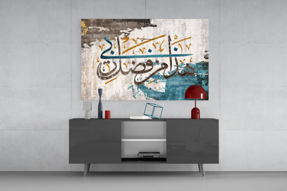 Modern living room interior with glass wall art displaying Arabic calligraphy in teal, gold, and brown tones mounted above gray media console.