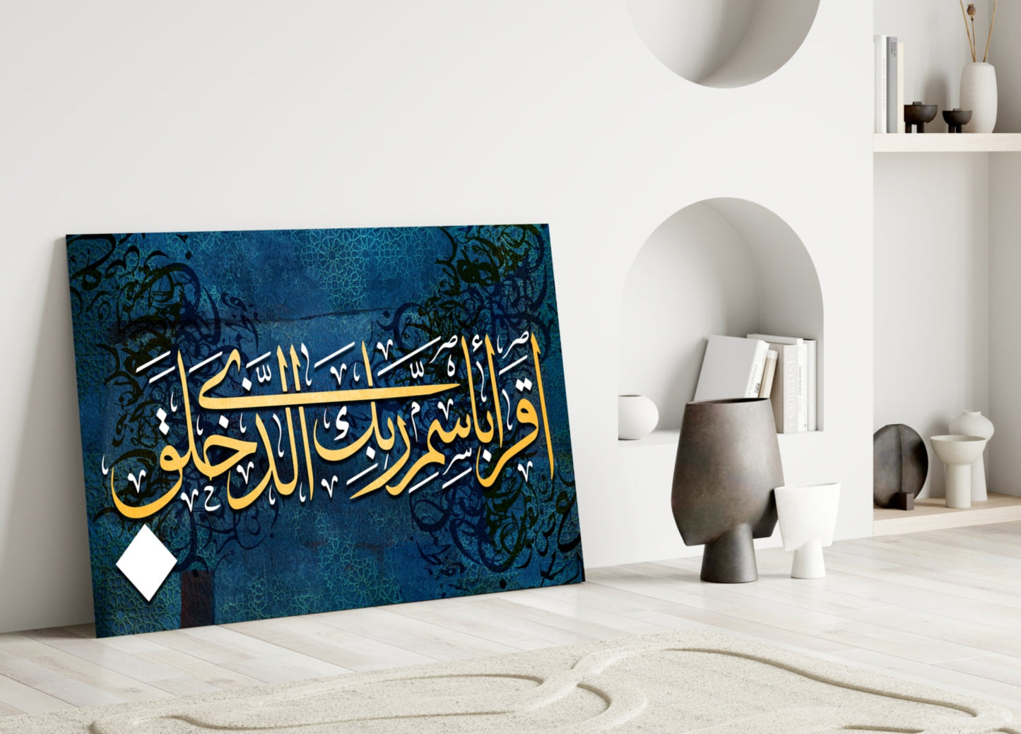 Surah Alaq glass wall art leaning against white wall in minimalist room with blue background, gold Arabic calligraphy, and decorative black patterns.