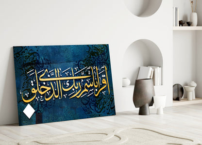 Surah Alaq glass wall art leaning against white wall in minimalist room with blue background, gold Arabic calligraphy, and decorative black patterns.