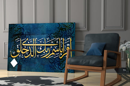 Surah Alaq glass wall art with gold and white Arabic calligraphy on deep blue patterned background, displayed in modern interior with wooden chair.
