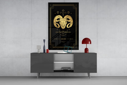 Black glass wall art featuring gold Aries zodiac ram head with decorative borders, displayed above gray media console in modern interior setting.