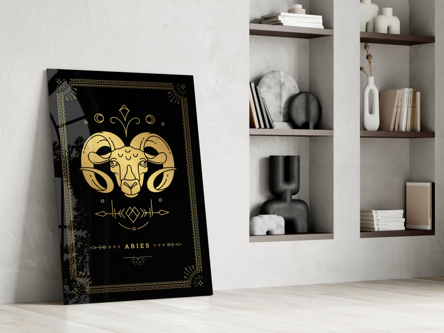 Black glass Aries zodiac art leaning against white wall beside white shelving with decorative objects and books.