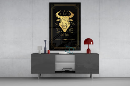 Black glass wall art featuring gold Taurus zodiac bull design displayed above gray console table with red lamp and decorative accessories.